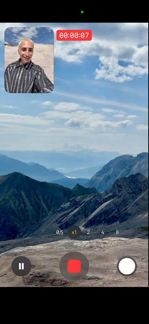 Una persona muestra la prestación Captura Dual grabándose con la cámara frontal mientras graba con la cámara trasera un paisaje con unas montañas