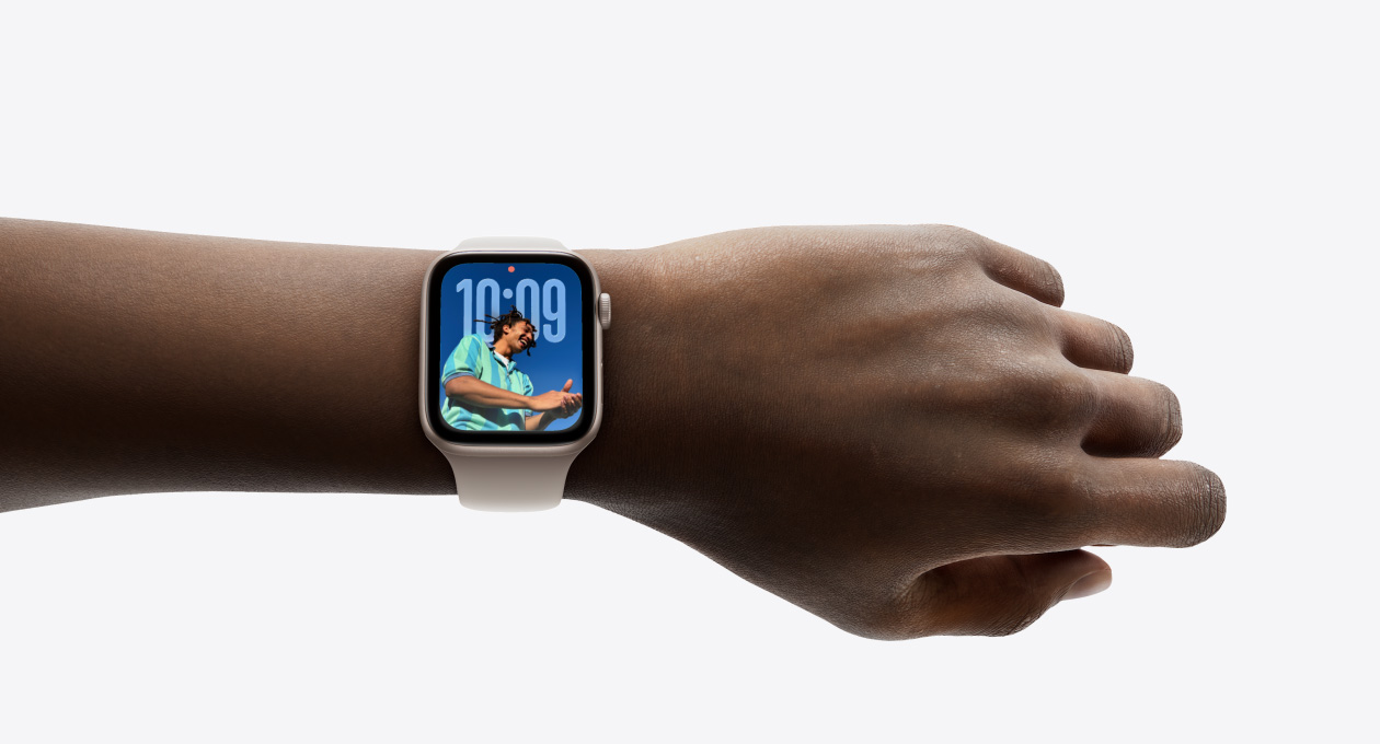 Bir kişinin bileğindeki kauçuk kordonlu Gümüş rengi Apple Watch’un yakından çekilmiş görseli, kadranda yeni Liquid Glass tasarımıyla Saat uygulaması görülüyor